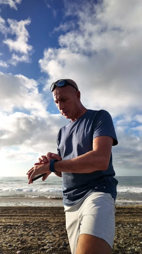 Mature man synchronizing his smartphone with smartwatch on the beach Stock Footage 346177802
