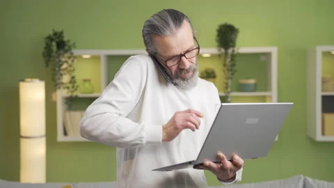 Mature man talks on the phone using a laptop at home. Business speech, meeting,  Video stock 231006993