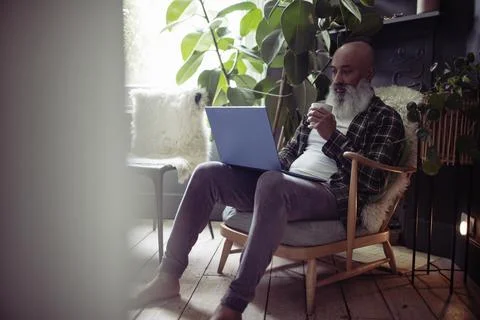 Mature man with tea using laptop in armchair at home 스톡 사진