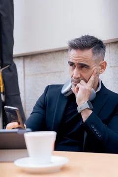 Mature man thinking while checking smartphone with tablet and coffee on table Stock Photos