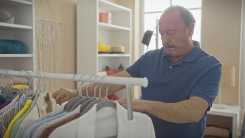 A mature man thoughtfully selects clothes from a rack in a brightly lit, we.. Stock Footage 277865160