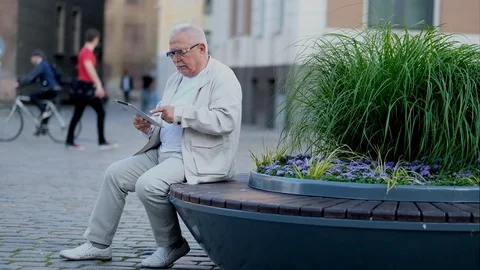 Mature man uses tablet resting on bench at flowerbed Stock Footage 115884257