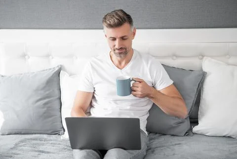 Mature man using laptop in bed with coffee Foto stock