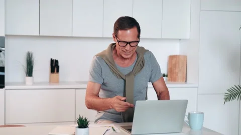 Mature man using laptop, making video call, sitting at table in kitchen, senior Stock Footage 231706482