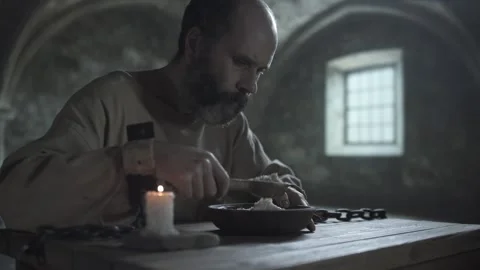 Mature medieval man in chains eating gruel with a spoon from a wooden bowl Video stock 337649691