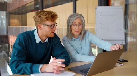 Mature mentor teaching intern, explaining online job using laptop in office. Stock Photos