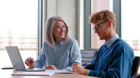 Mature mentor teaching intern, explaining online job using laptop in office. Stock Photos