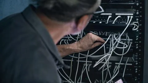Mature specialist inserts cables into server rack, uses tablet computer Stock Photos