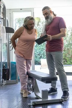 Mature trainer showing tablet while senior woman leaning near flat bench in gym Stock Photos