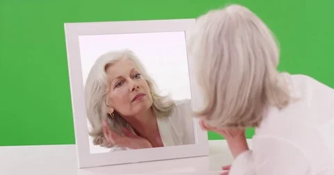 Mature woman in her 50s checking her reflection in the mirror on greenscreen Stock Footage