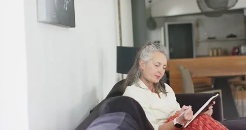 Mature woman settling on couch, pulling up throw, tapping tablet with stylus to Stock Footage 341851811