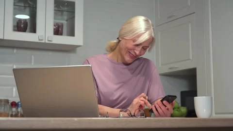Mature woman smiling while using smartphone and laptop in modern kitchen setting Stock Footage 308442484