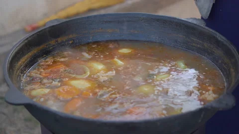 Meat and vegetable soup is cooked in a large cast-iron cauldron over an open Stock Footage 156410314