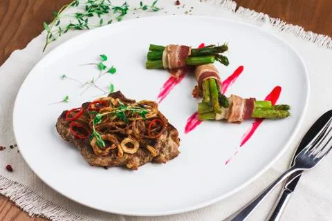 Meat steak under fried onions and peppers with asparagus and bacon on a white Stock Photos
