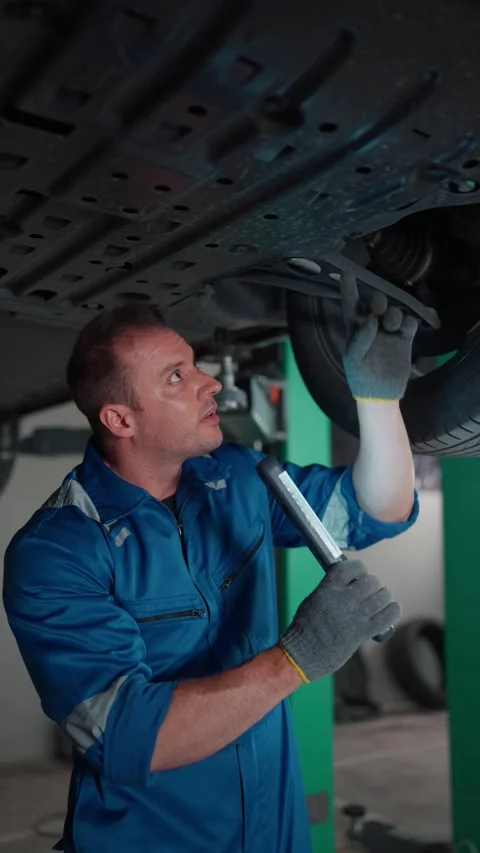 Mechanic inspects car underside with flashlight and tool Stockbeeldmateriaal 347769759
