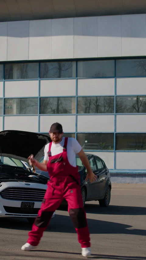 Mechanic in red overalls humorously dancing outside workshop with car hood open Stock Footage 320394239