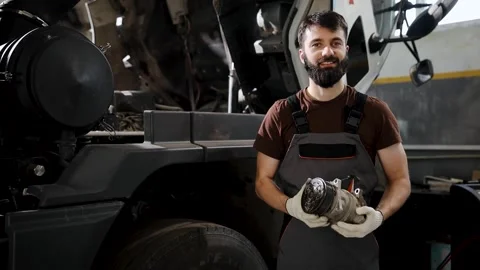 Mechanic smiling, holding engine part, working on truck in auto service garage 스톡 동영상 333705840