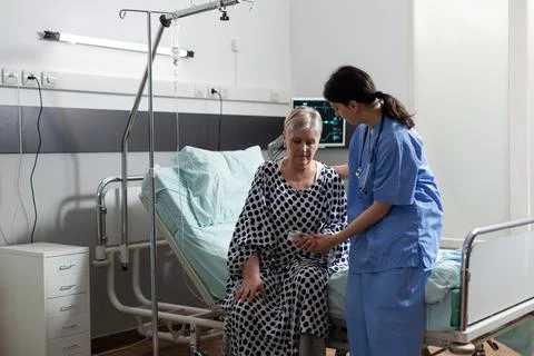 Medical nurse in scrubs helping senior woman in hospital room Stock Photos