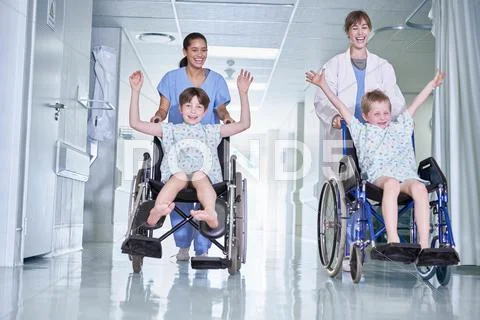 Medical Orderlies Having Fun Pushing Boy Patients In Wheelchair On Hospital