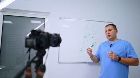 Medical presenter explaining a kidney diagram on camera. A man in blue scrubs Stock Footage 346615136