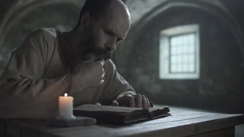 Medieval prisoner stroking beard and reading a book inside a gloomy dungeon Stock Footage 340079697