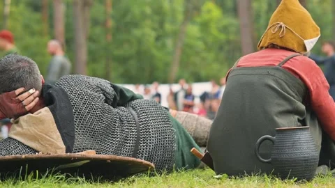 Medieval reenactors relax on the grass, dressed in authentic clothing typical of Видео 345032922