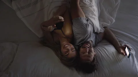 Medium elevated view of happy couple lying in bed with smart phones in their Stock Footage 277968410
