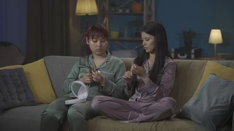 Medium-full video capturing two young women wearing pajamas sitting on the couch Stock Footage 252228629