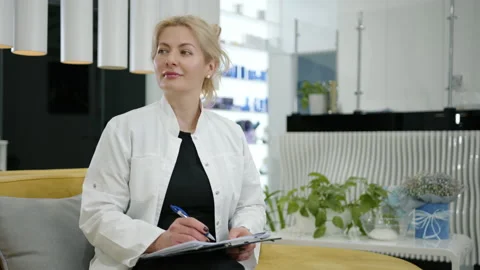 Medium shot of cute blonde in lab coat, sitting on sofa, making notes in clinic Stock Footage 239863311