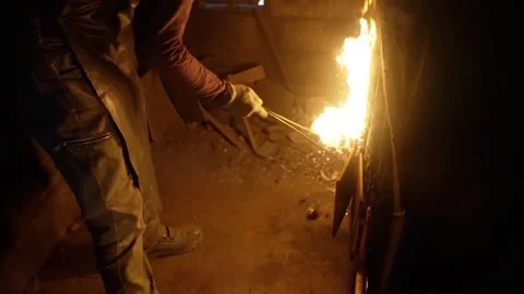 Medium shot of a foundry worker in protective apron using long tongs to stoke a Stock Footage 332376463