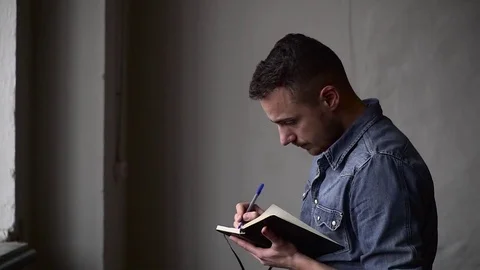 Medium shot of the guy writes in the notebook while standing near the window Video stock 82904664