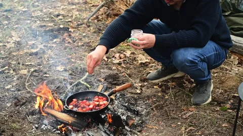 Medium shot of a man cooking cherry tomatoes in a frying pan over a campfire Stockbeeldmateriaal 334681107