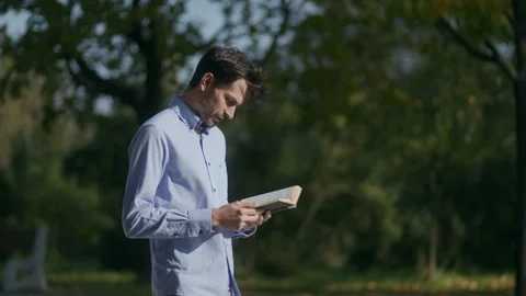 Medium shot of a man standing in the park and reading a black book Video stock 332299080