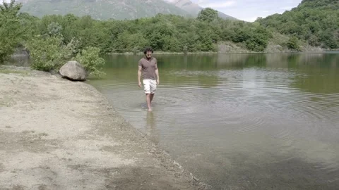 Medium shot Man walks with his feet in the water on the shore of a lake. 스톡 동영상 154606933