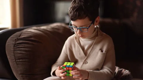 Medium shot of smart child boy wearing eyeglasses solving Rubik Cube and talkign Video stock 165395147