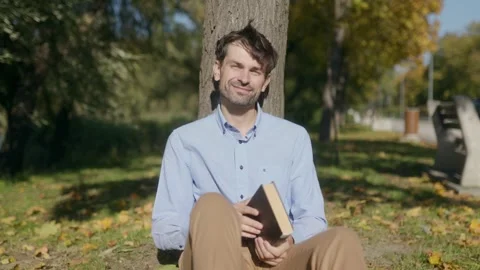 Medium shot of a smiling man closing a book while sitting under a tree Stock Footage 332298966