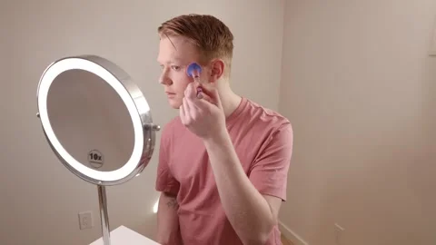 A Medium Shot of A Young Man Applying Blush In Front Of A Ring Light Mirror Stock Footage 309892880