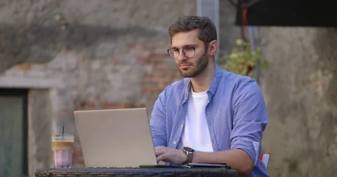 Medium shot of young man in blue shirt sitting at table in outdoor cafe or Stock Footage 166298638