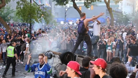 Men atop upside down car cheer and take photos during Vancouver 2011 riots Stock Footage 127201001