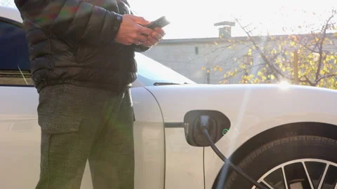Men in black jacket charges a white electric car on the street from charging Stock Footage 233604316