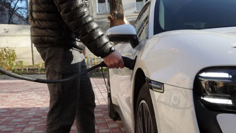 Men in black jacket charges a white electric car on the street from charging Stock Footage 233604424