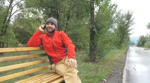 Men guy in the cap talking on phone sits on bench in autumn in city park. 库存影片 68684326