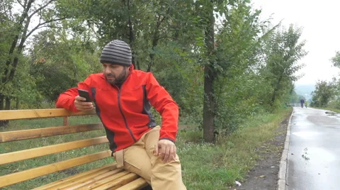 Men guy using app on smartphone sits on bench in autumn in city park. 스톡 동영상 67490801