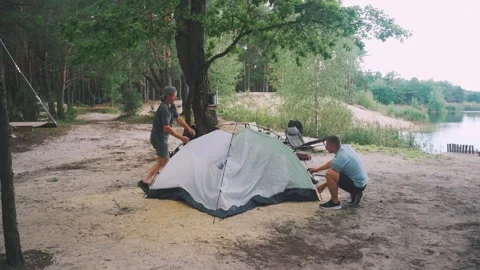 Men organize camping, Two individuals set up tent near water, Two men assemble Stock Footage 323290697