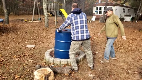 Men place blue empty barrel upside down onto fire pit to burn it inside Stock Footage 236170919