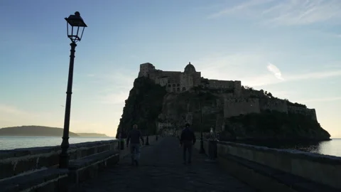 Men walk on rock bridge towards ancient Italian coastal castle Stock Footage 220181836