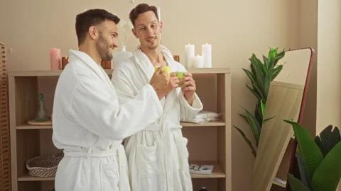 Men wearing bathrobes drinking tea in spa room featuring candles plants and.. Stock Footage 289700527
