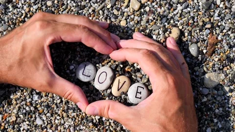 Men's hands folded in shape of a heart hold sea stones with word love written Stock Footage 138748615