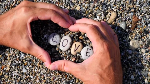 Men's hands folded in shape of a heart hold sea stones with word love written Stock Footage 138751901