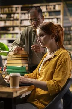 Mentor stands beside student using laptop at library table with books Foto stock